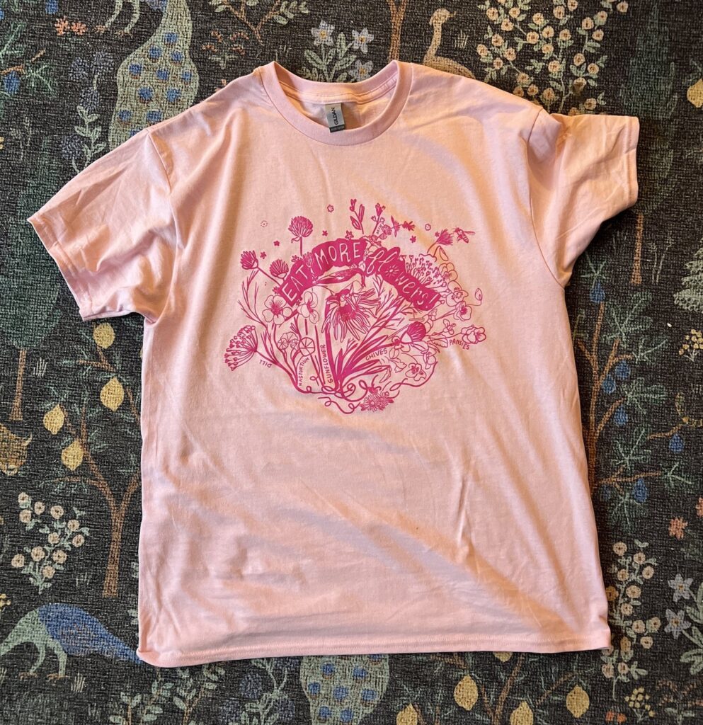 A pink t-shirt illustrated with a screen print of flowers and the text "EAT MORE flowers!"