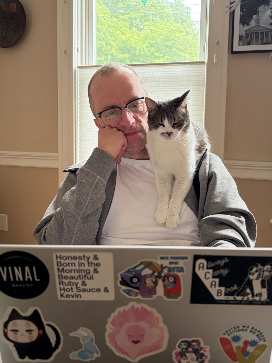 Ethan leaning on one hand and regarding an open laptop with a cat perched on his shoulder.