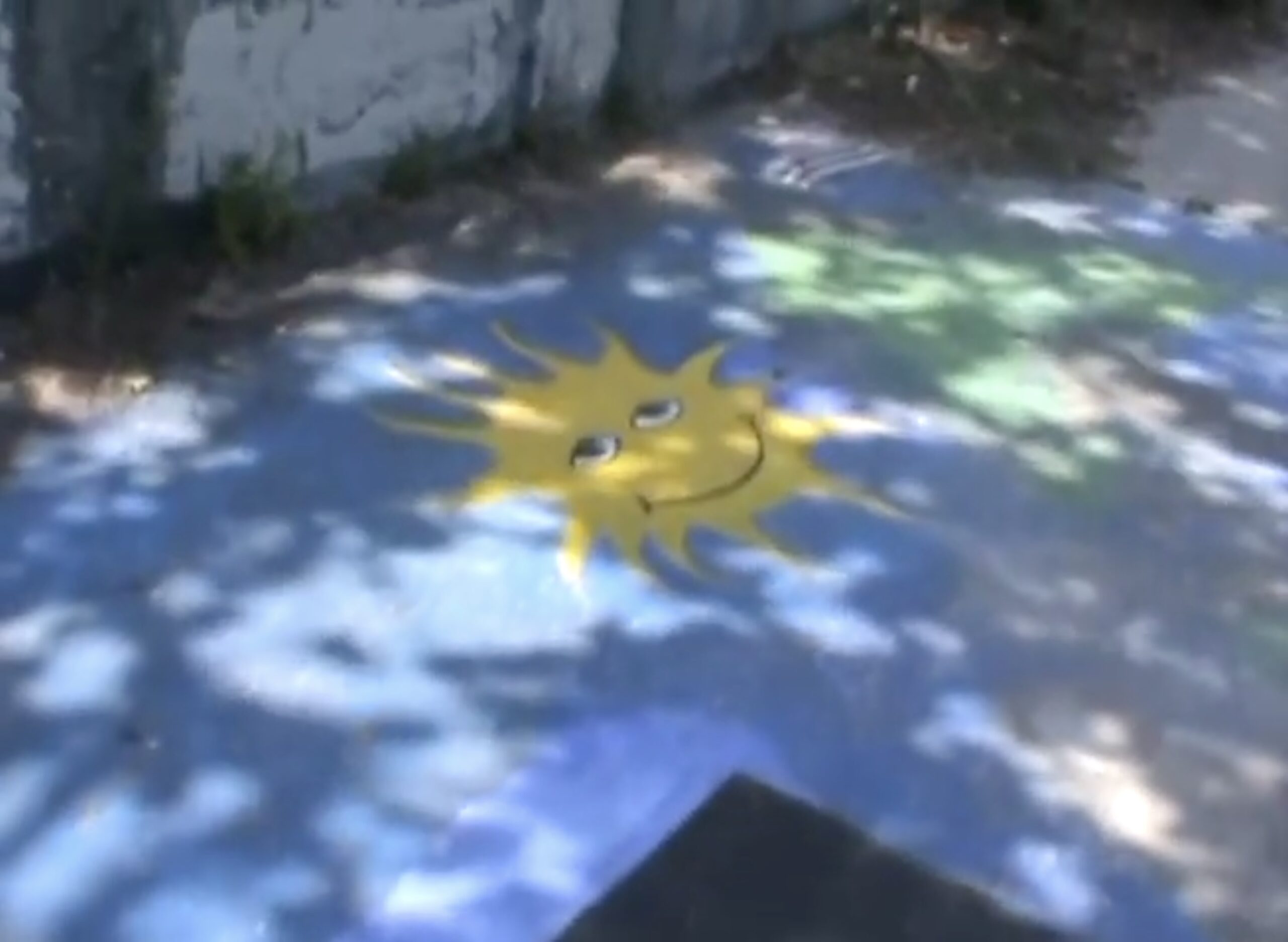 Screenshot of a low-res digital video, with dappled shade on a sun-face painted on concrete.
